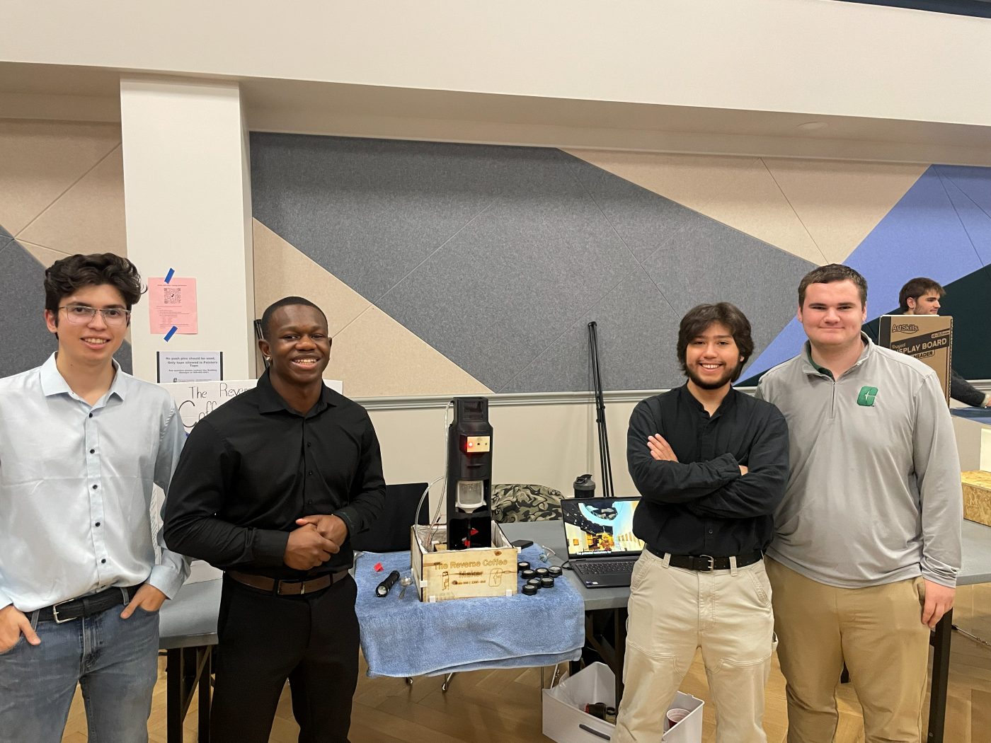 Four people stand around a wooden-box prototype labeled The Reverse Coffee Maker on an expo table